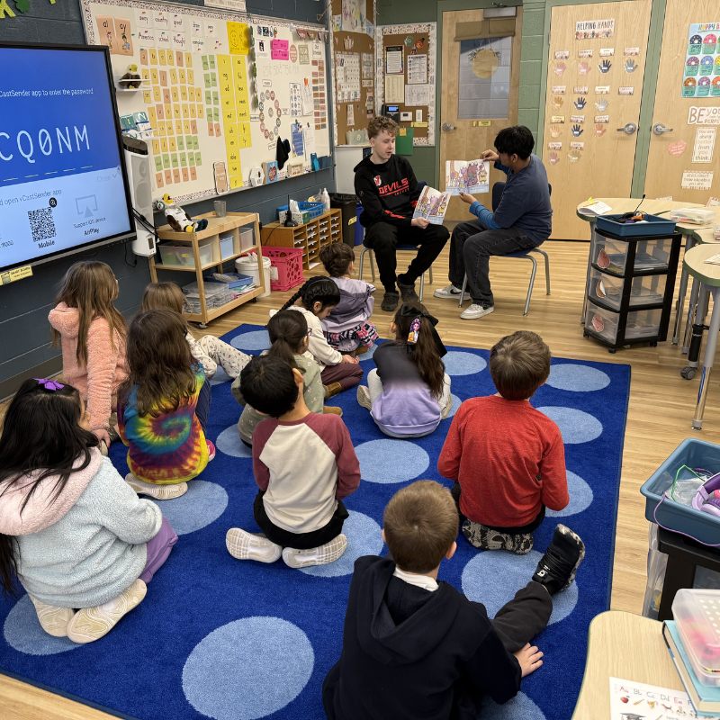 Two teenagers read a book aloud to a group of young students sitting on a blue rug in a classroom.