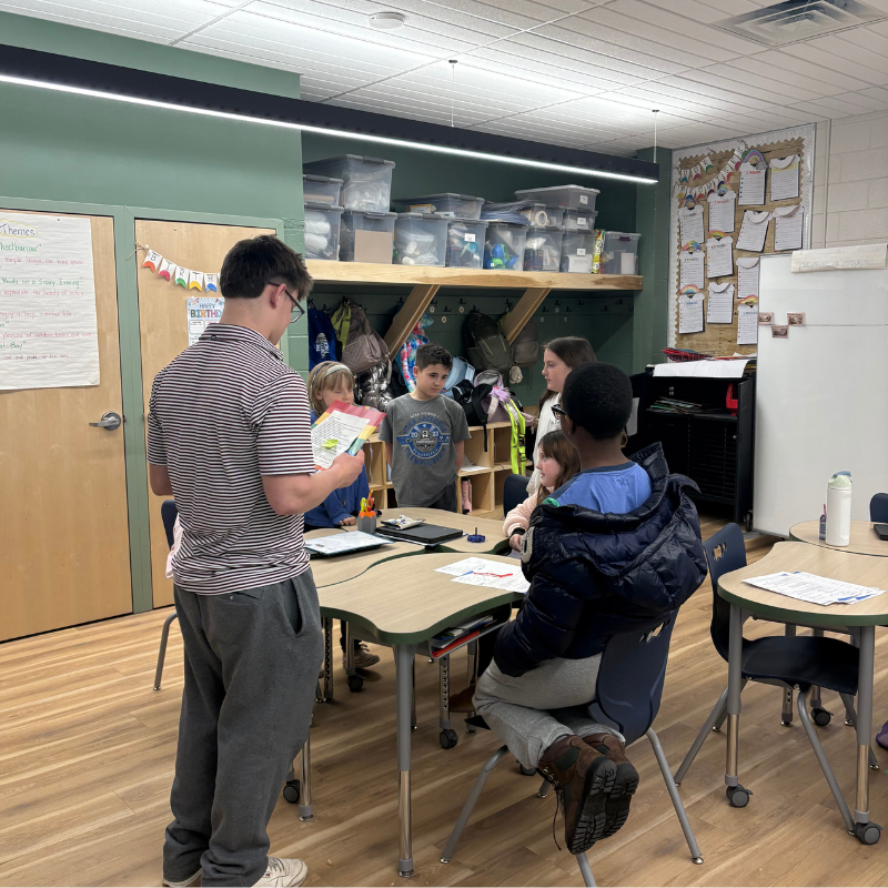 A group of students gather around a table in a classroom, listening to a young man holding a paper.