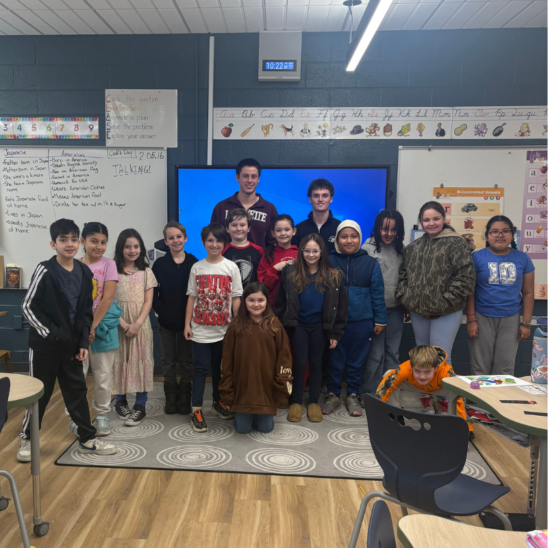 A group of students and two young men stand together in a classroom in front of a large screen.