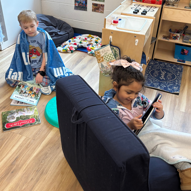 A young boy wrapped in a blue blanket reads books on the floor, while a girl in the foreground looks at a tablet.