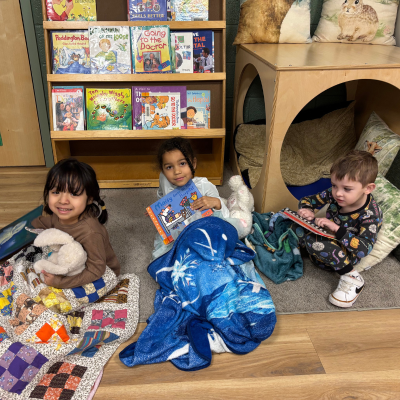 Three young children sit on the floor with blankets and books in a room with bookshelves.