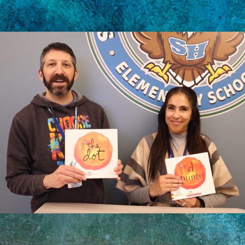 A man and a woman stand in front of a school logo, each holding a children's book.
