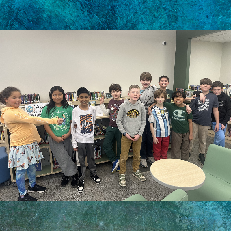 A diverse group of elementary school children stand together in a library, smiling.