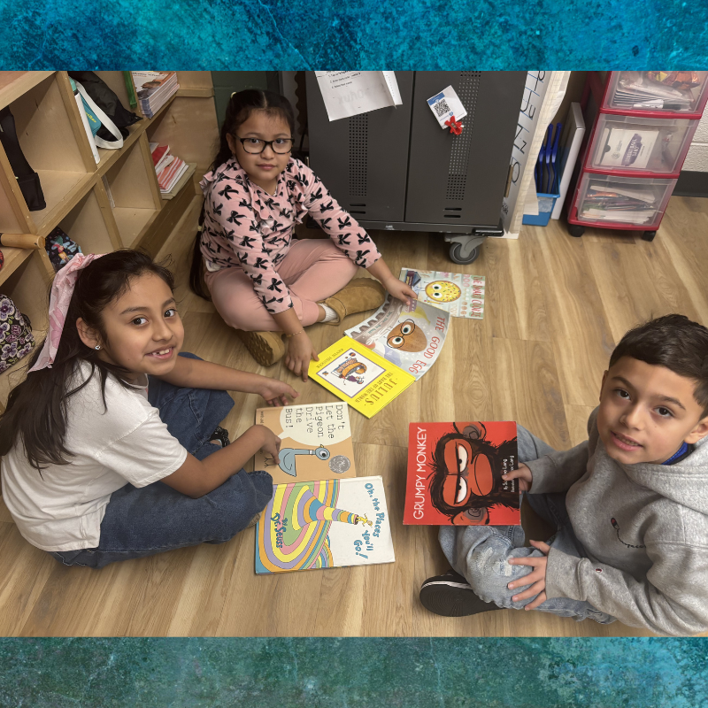 Three children sit on a classroom floor surrounded by colorful children's books.
