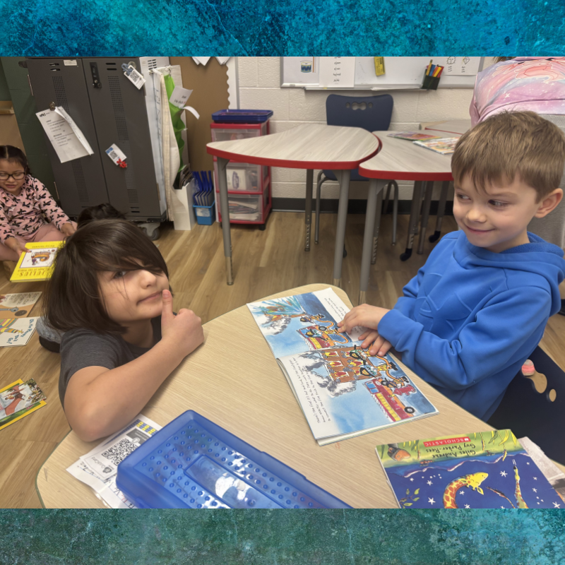Two children sit at desks in a classroom, looking at open books.