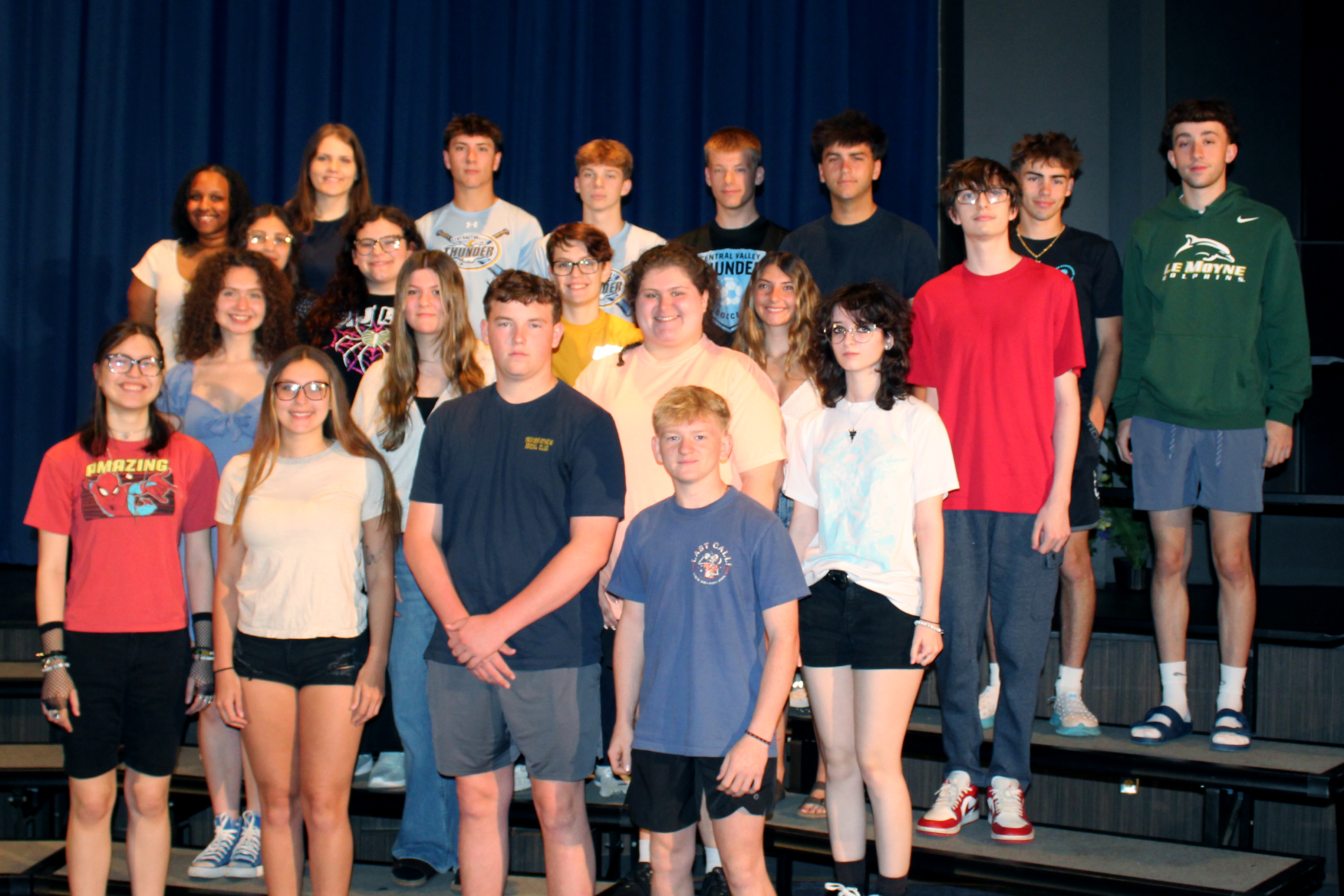 A diverse group of young people stand on risers in front of a blue curtain.