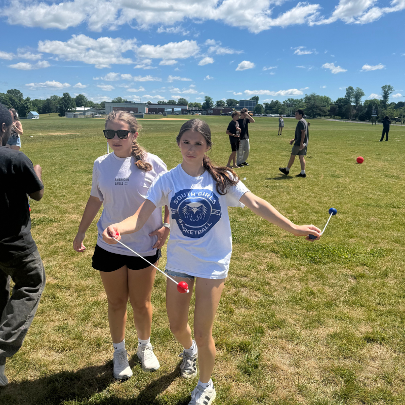 Two young women play a game with colorful balls on strings in a grassy field.