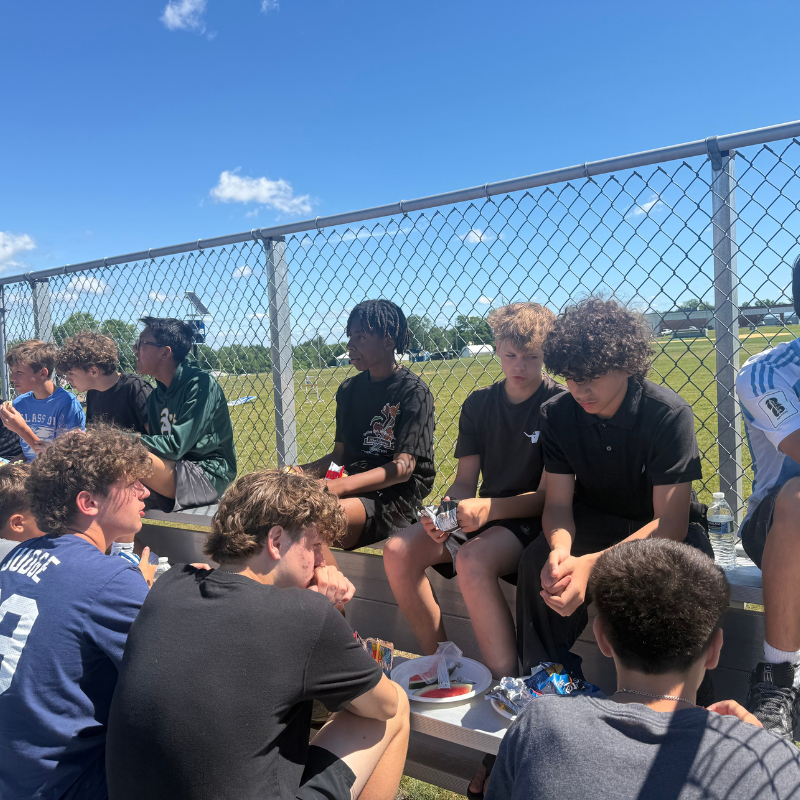 A group of young men sit on bleachers under a bright blue sky, taking a break.