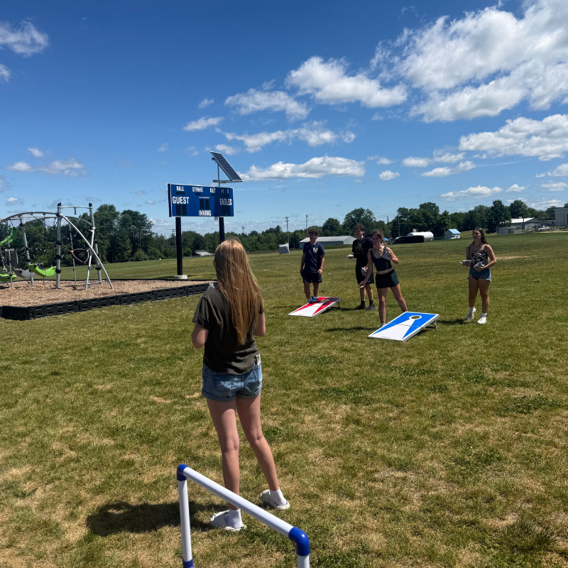 Teenagers play cornhole on a sunny day with a scoreboard in the background.