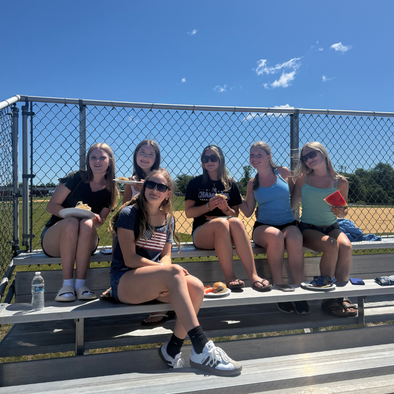 Six young women sit on bleachers, enjoying snacks and drinks on a sunny day.