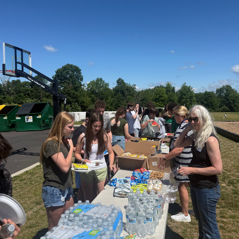 A group of people, mostly young adults, gather around a table laden with snacks and drinks outdoors.