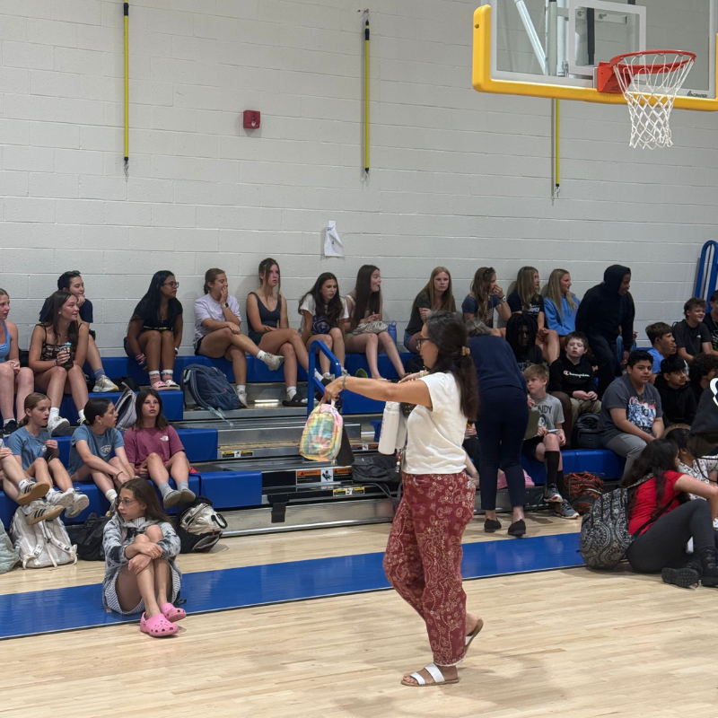 A group of young people sit on bleachers in a gymnasium, listening to a woman.