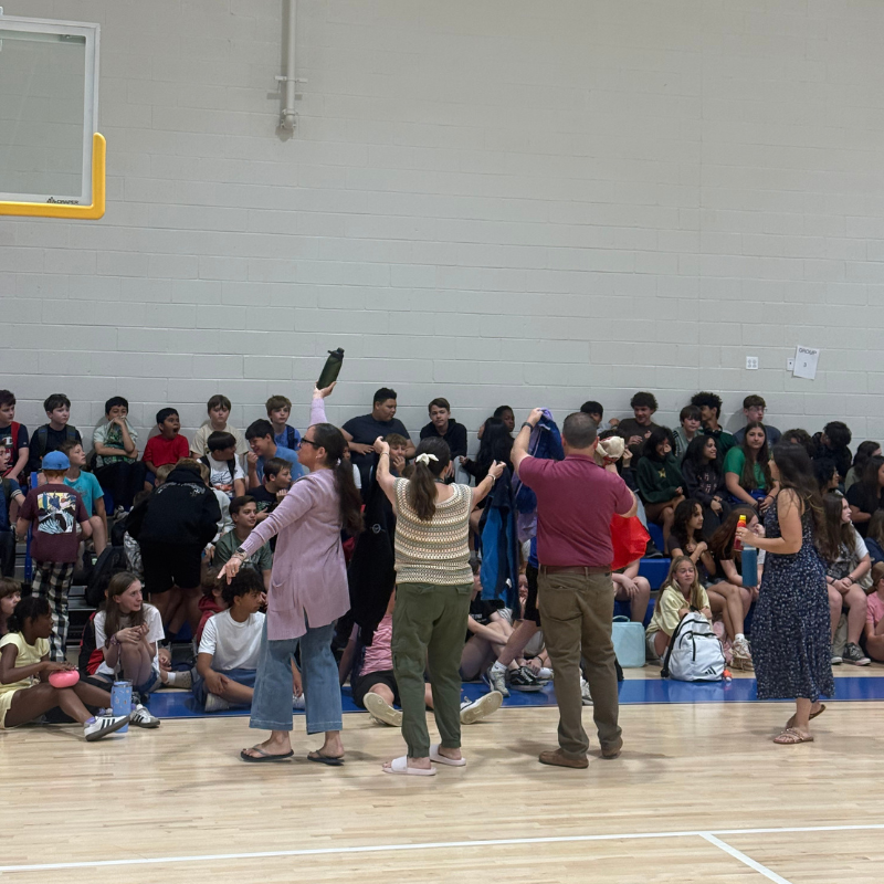 Adults interact with a seated audience of students in a gymnasium.