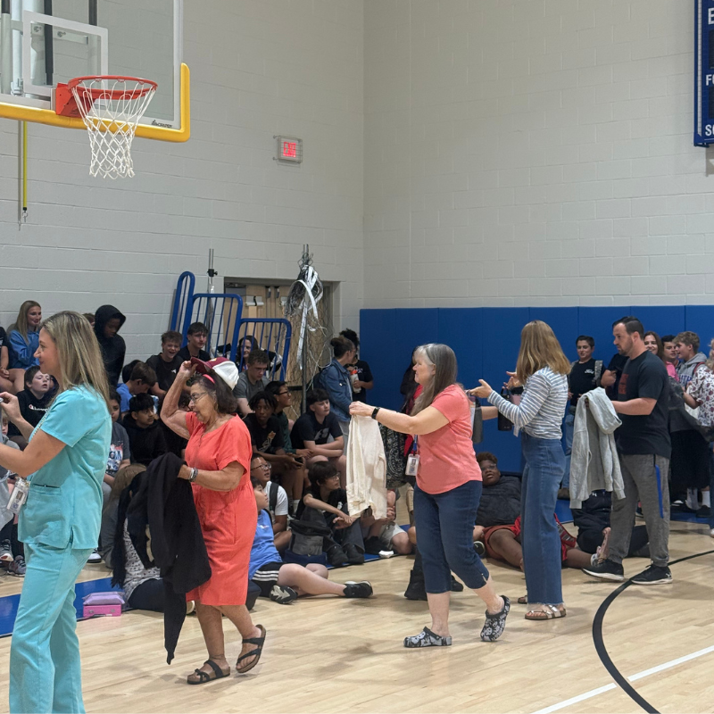 People gather in a gymnasium, with some seated on the floor and others standing.