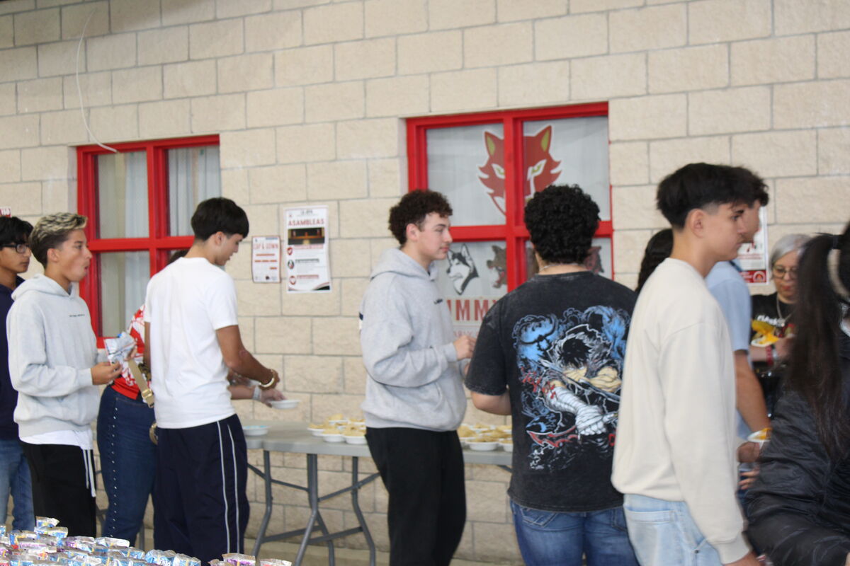A group of students stands in line at a table with food in front of a brick wall with red-trimmed windows.