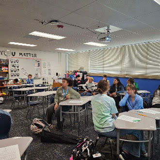 Students sit at desks in a brightly lit classroom, facing the front while listening to a lesson.