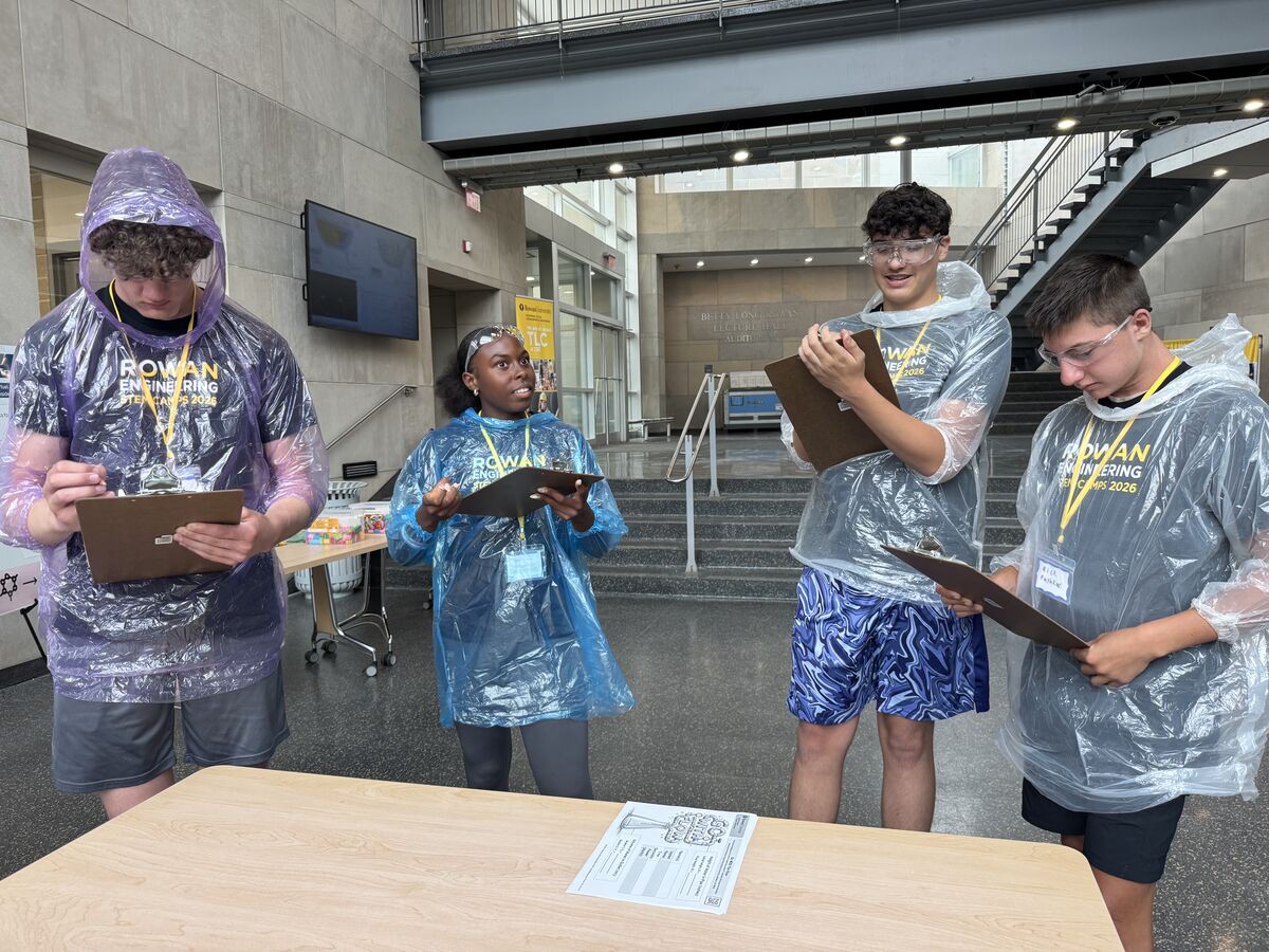 Four young people in clear ponchos stand at a table, holding clipboards and looking at papers.