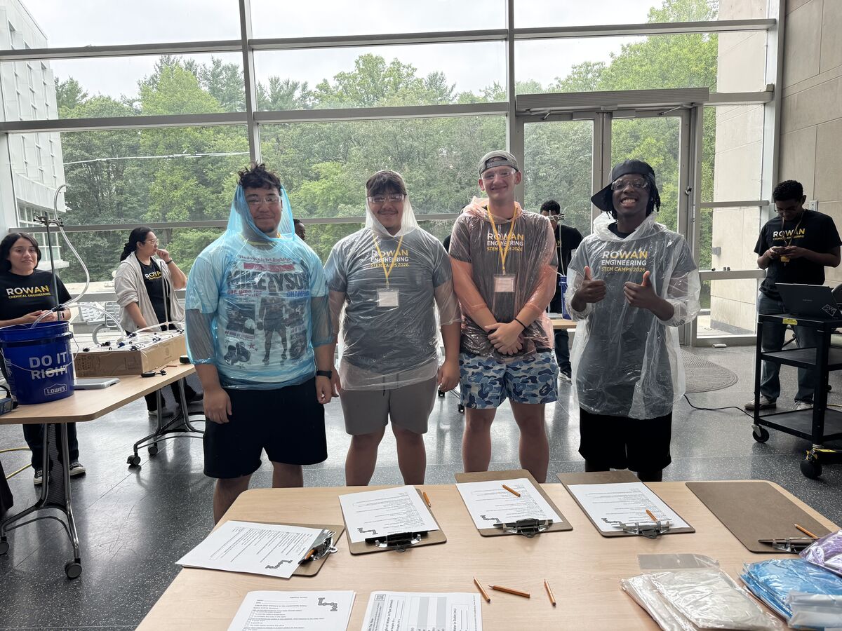 Four young people wearing clear ponchos stand behind a table with clipboards and papers, with a large window and greenery in the background.