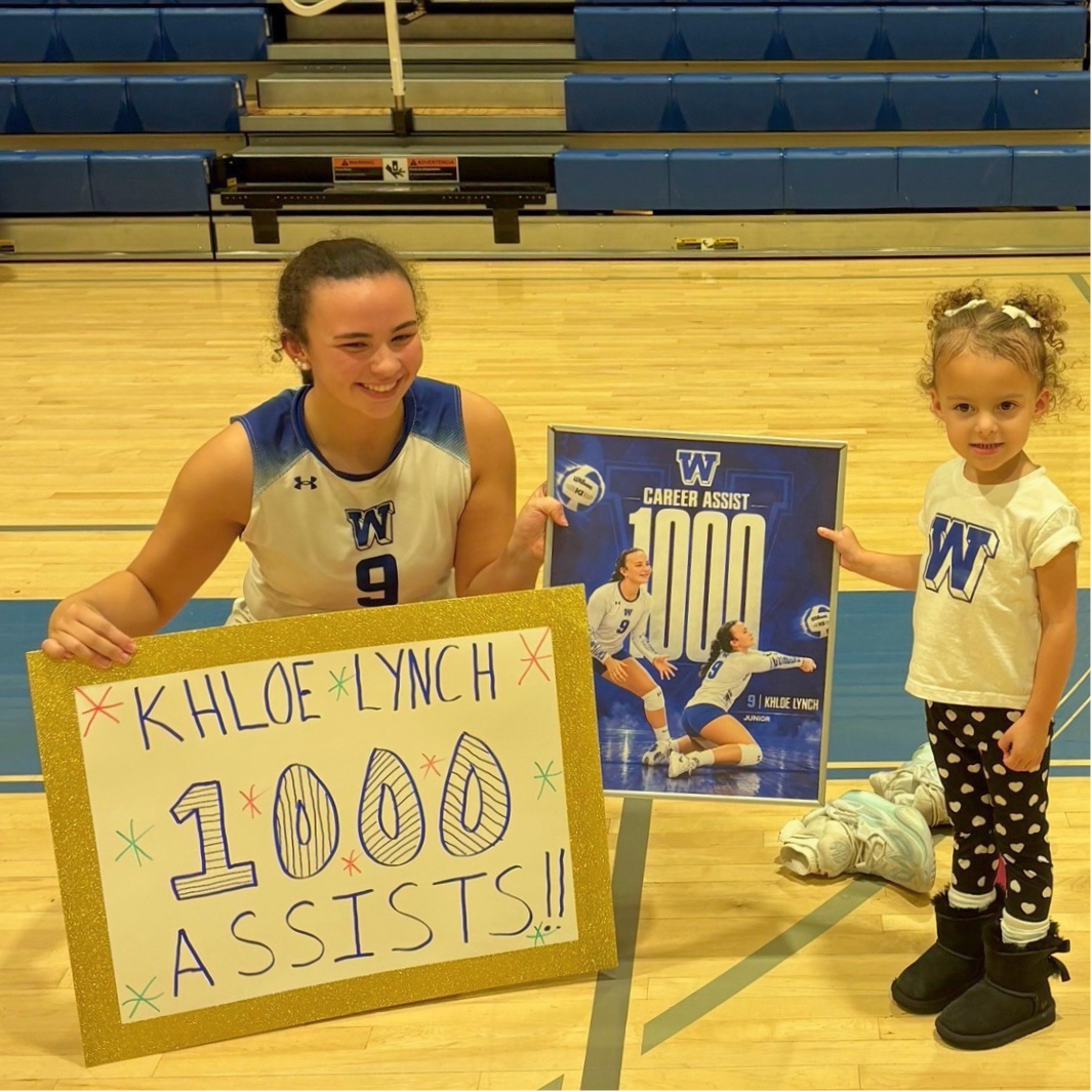 A smiling volleyball player in a white and blue jersey holds a handmade sign that reads "KHLOE LYNCH 1000 ASSISTS!!" while a young child holds a framed commemorative poster.