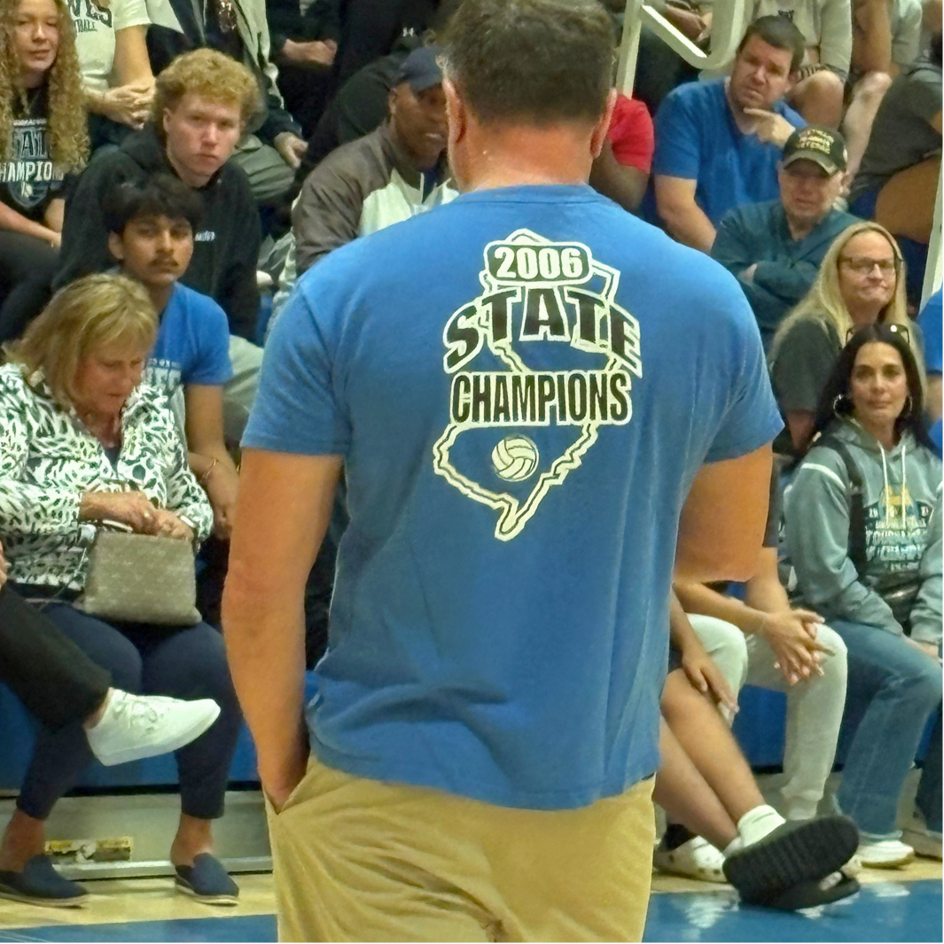 A person wearing a blue t-shirt that reads "2006 State Champions" stands before a seated crowd in a gymnasium.