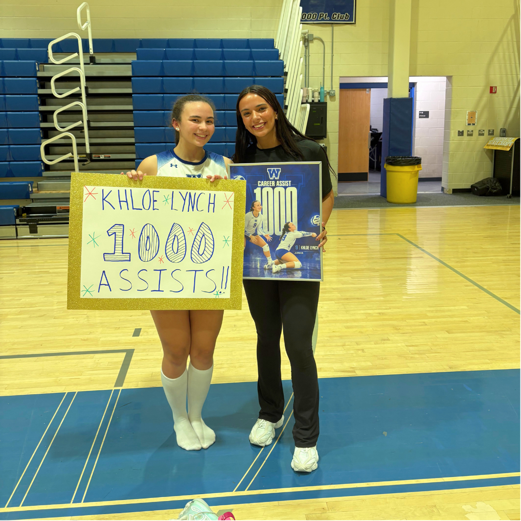 Two young women smile on a gym basketball court, holding a large sign that reads "KHLOE LYNCH 1000 ASSISTS!!" and a framed commemorative photo.