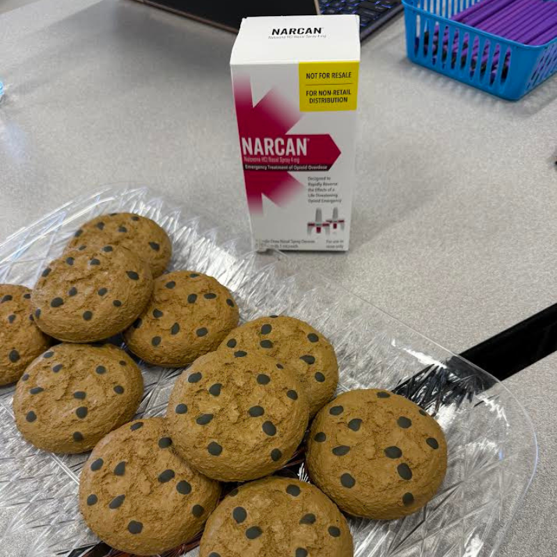 A clear tray holds several chocolate chip cookies next to a box of NARCAN nasal spray.