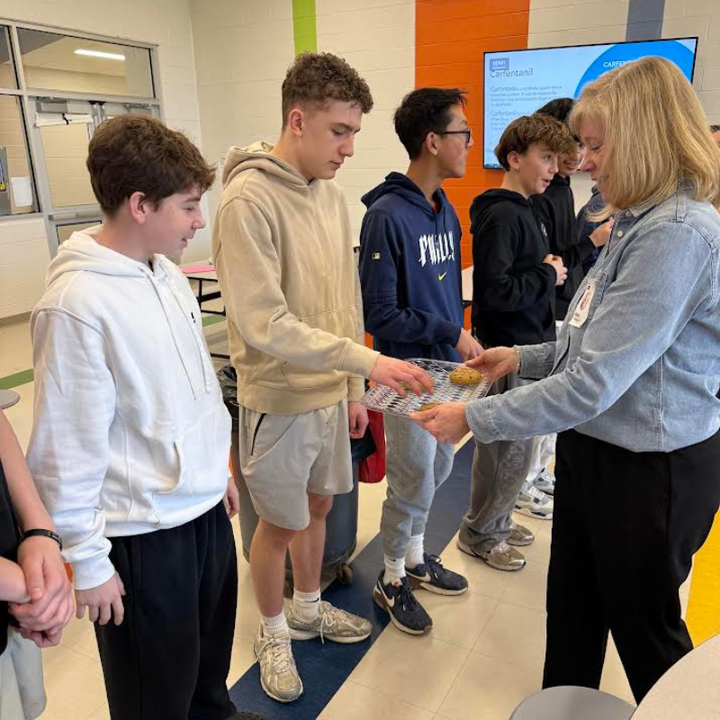 A woman offers cookies on a cooling rack to a group of young men.