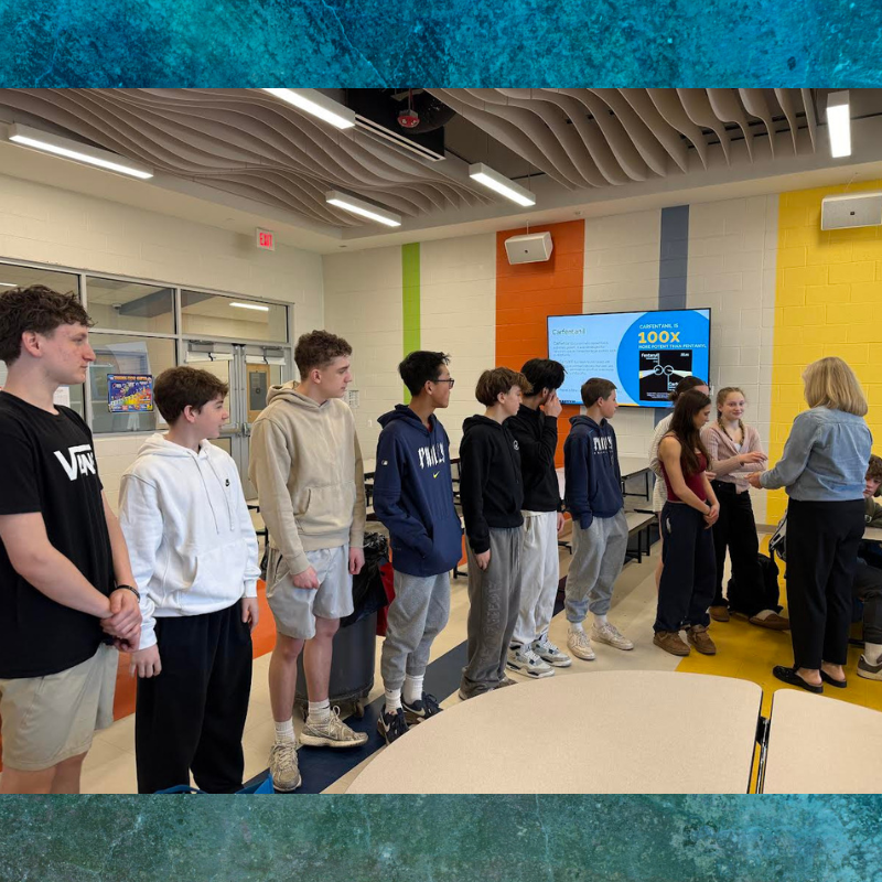 A group of students stands in a classroom, listening to a teacher.