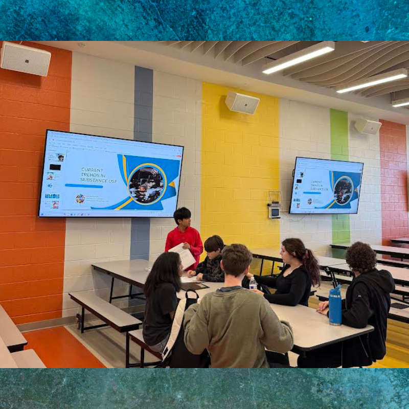 Students gather around tables in a brightly colored room, watching a presentation on a large screen.