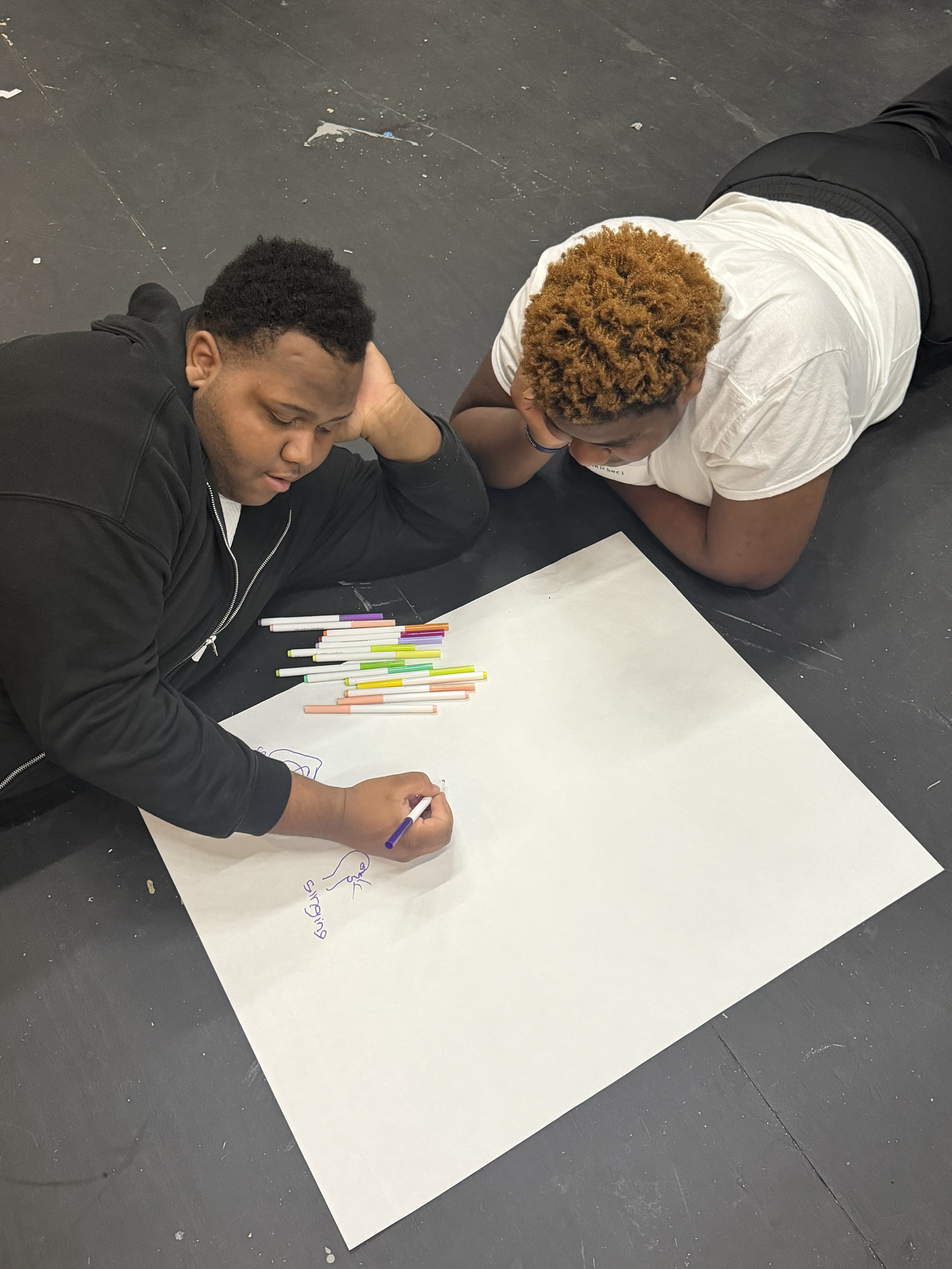 Two people lie on the floor, collaborating on a drawing on a large sheet of white paper with colored markers.
