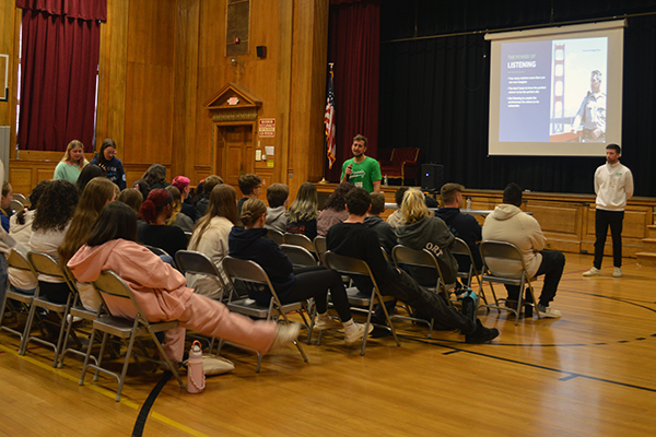 A presenter in a green shirt speaks to an audience seated in chairs in a gymnasium.