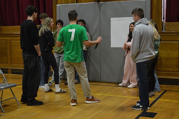 A group of young people stand in a gymnasium, listening to a speaker in a green shirt.