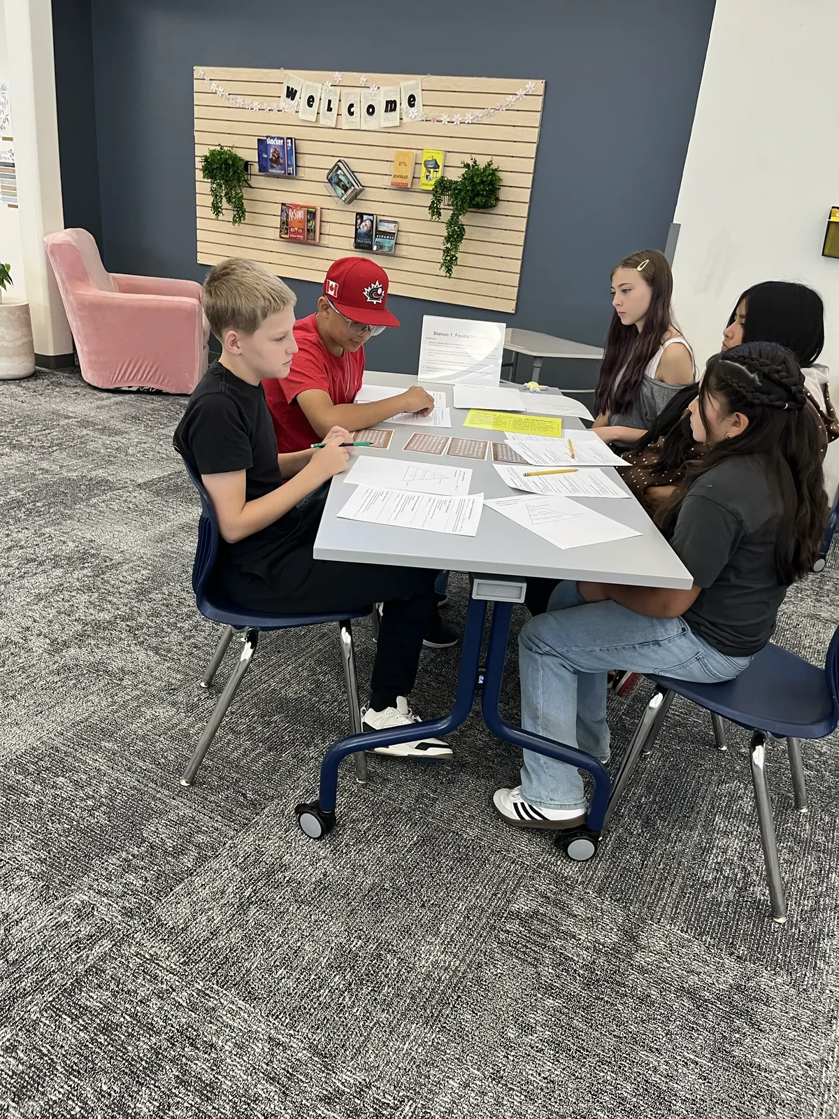 Students collaborate around a table, reviewing papers and worksheets in a classroom setting.
