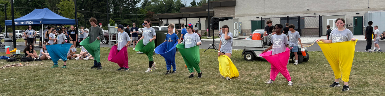 Students participate in a sack race event outdoors, wearing colorful fabric sacks.