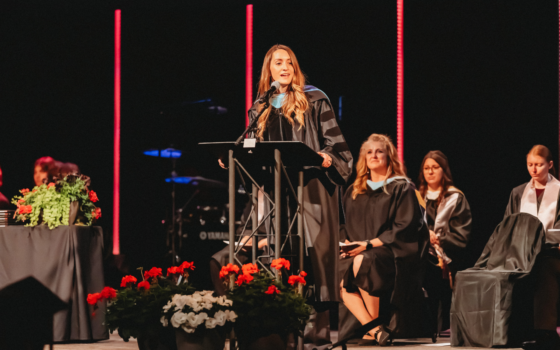 A woman in a graduation gown speaks at a podium on a stage with red lights.