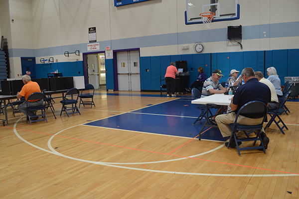 People are seated at tables in a gymnasium, with some interacting with a computer station.
