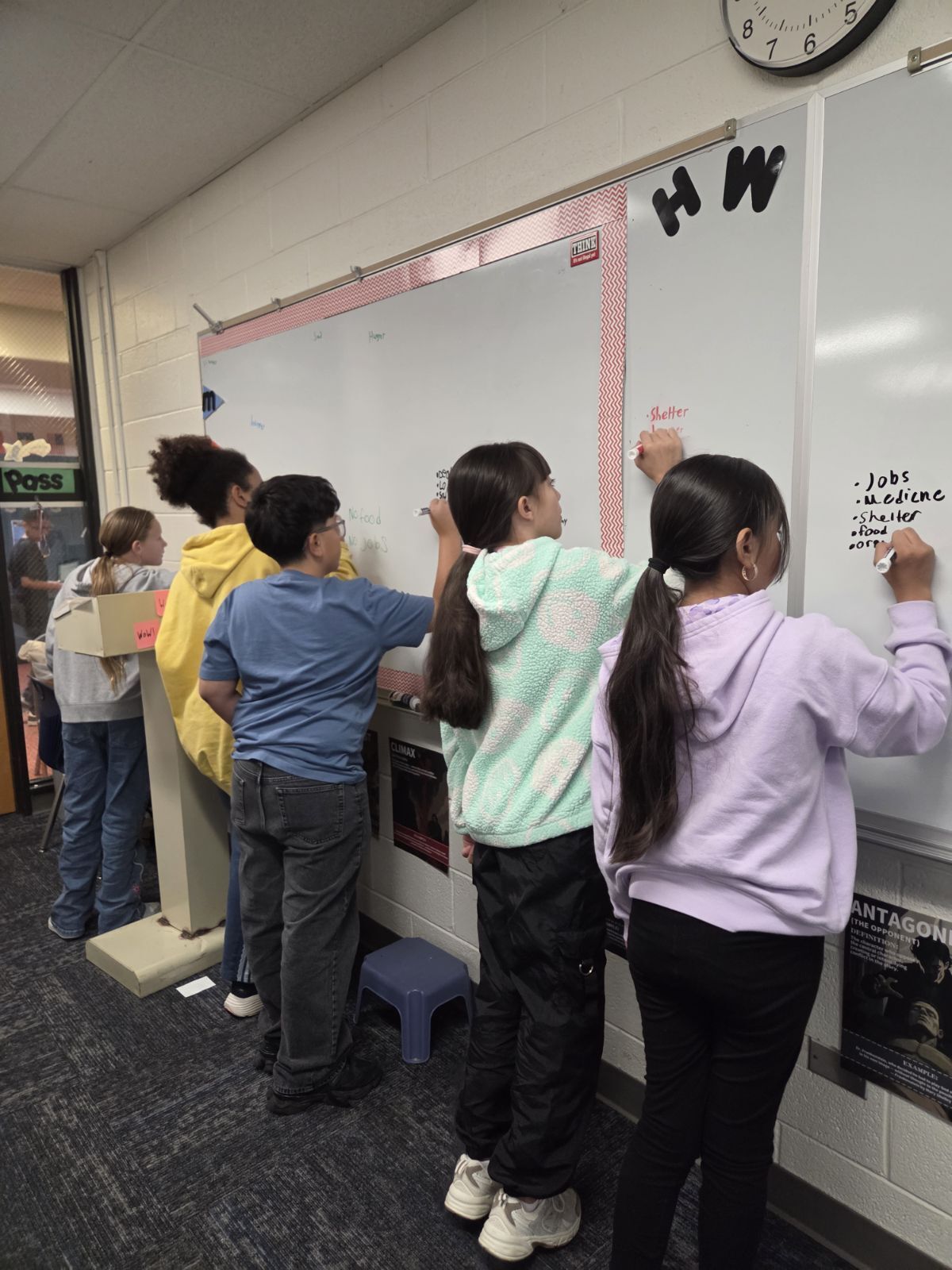 Five students stand in a line facing a large white board, each writing with a marker.