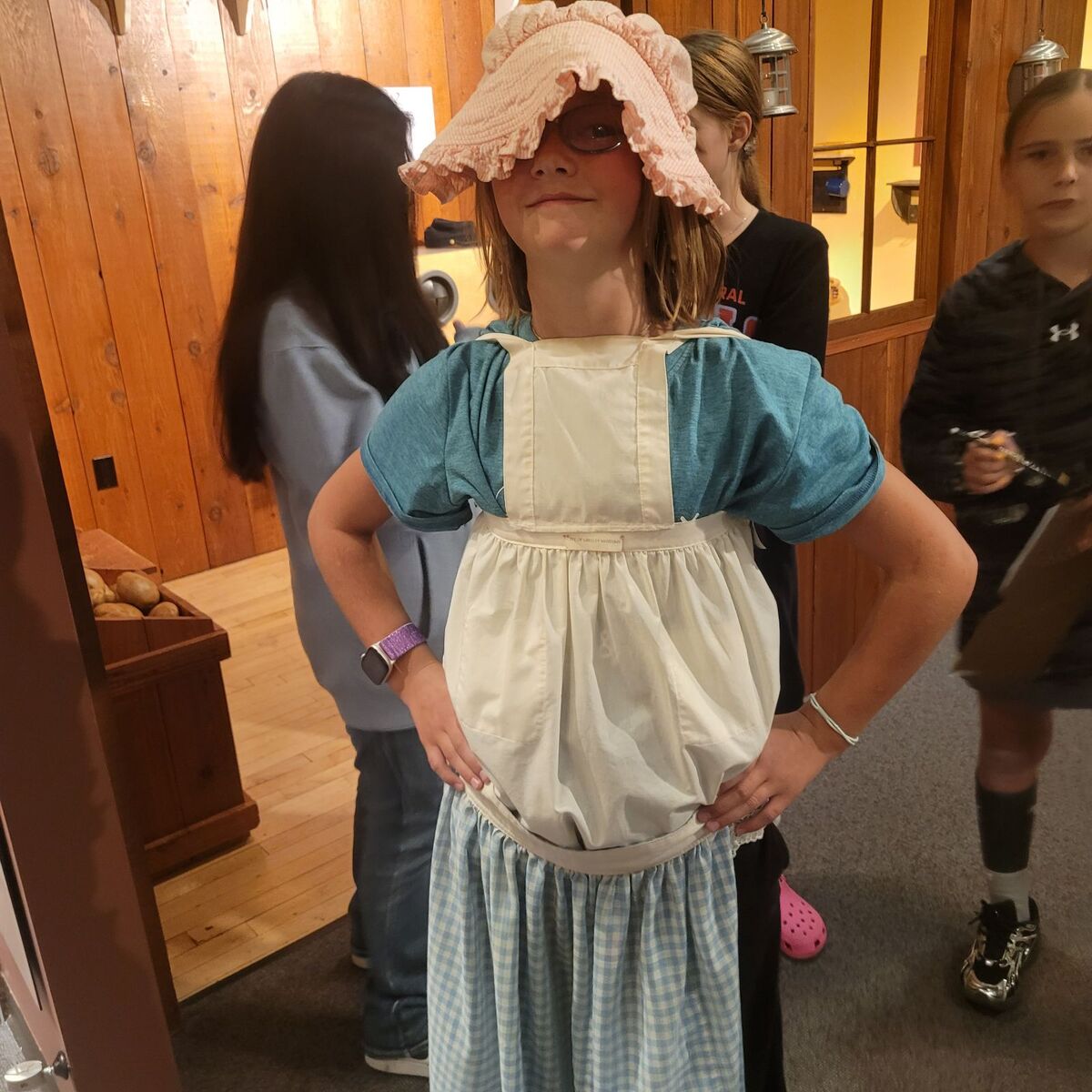 A child smiles while wearing a historical pioneer costume with a sunbonnet indoors.
