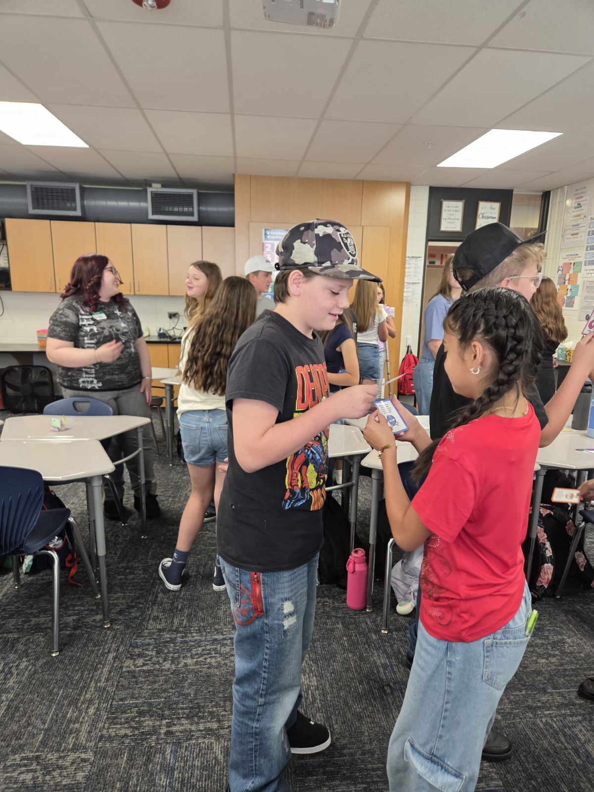 Students interact in a classroom, with two children in the foreground holding cards.