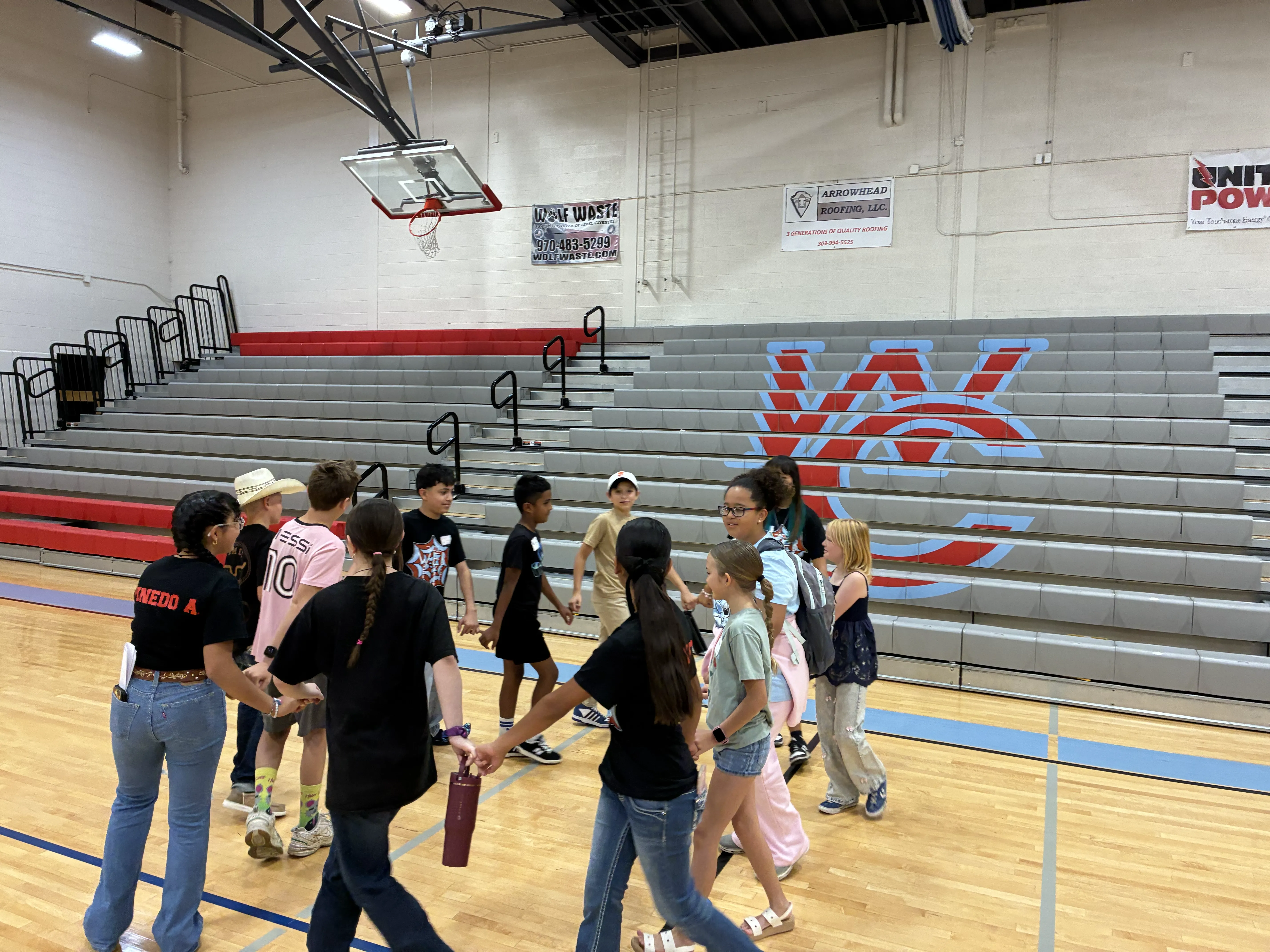 A group of children and adults hold hands in a circle on a basketball court, with bleachers in the background.