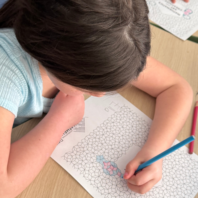 A young girl with dark hair colors a geometric pattern on a worksheet, with a blue pencil in her hand.
