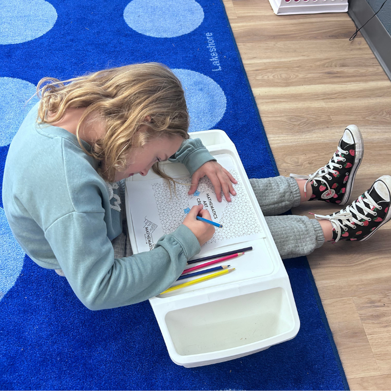 A young person with blonde hair colors a math worksheet on a portable lap desk.