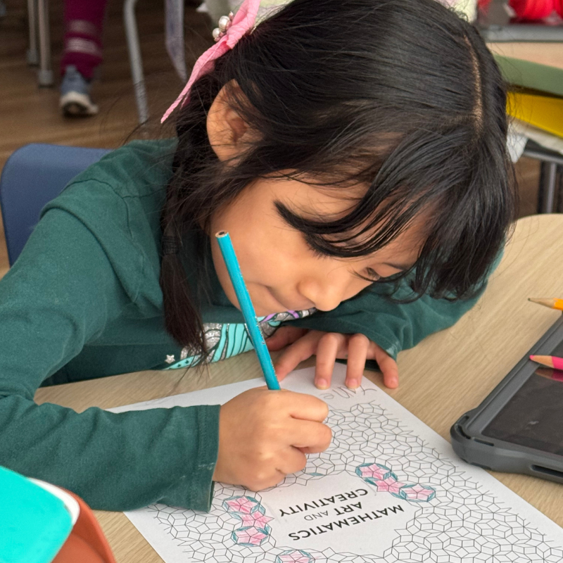 A young girl with a pink bow in her hair concentrates on coloring a geometric pattern on a worksheet.