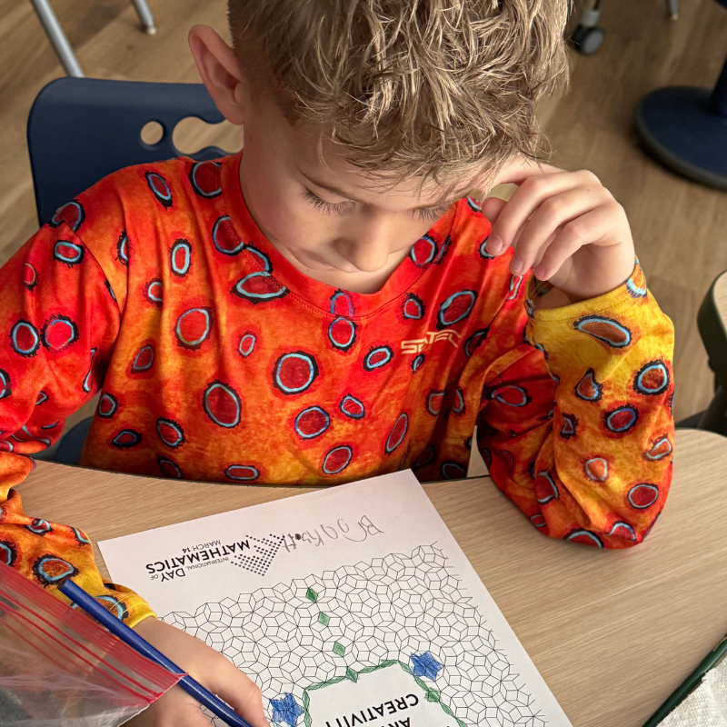 A young boy in an orange, patterned shirt focuses on a math activity sheet.