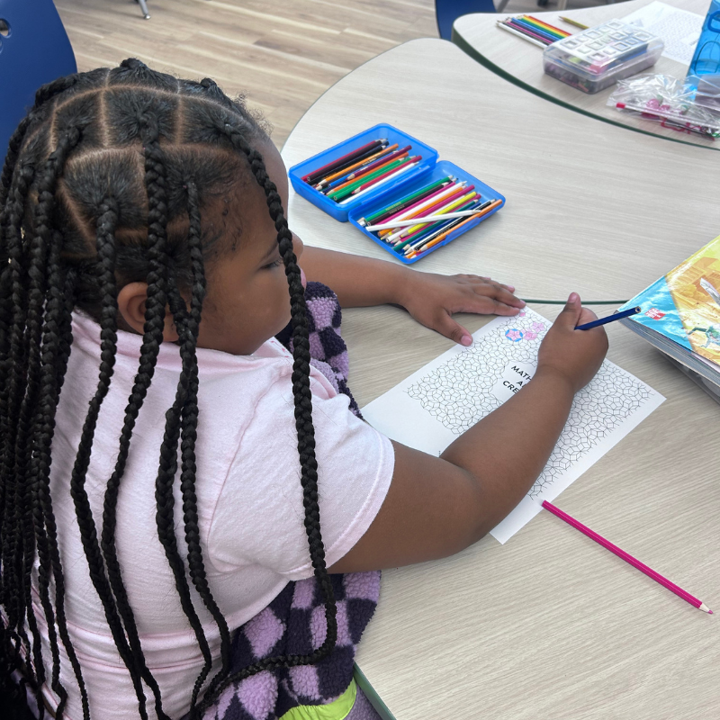 A young girl with long braids is coloring a worksheet at a table.