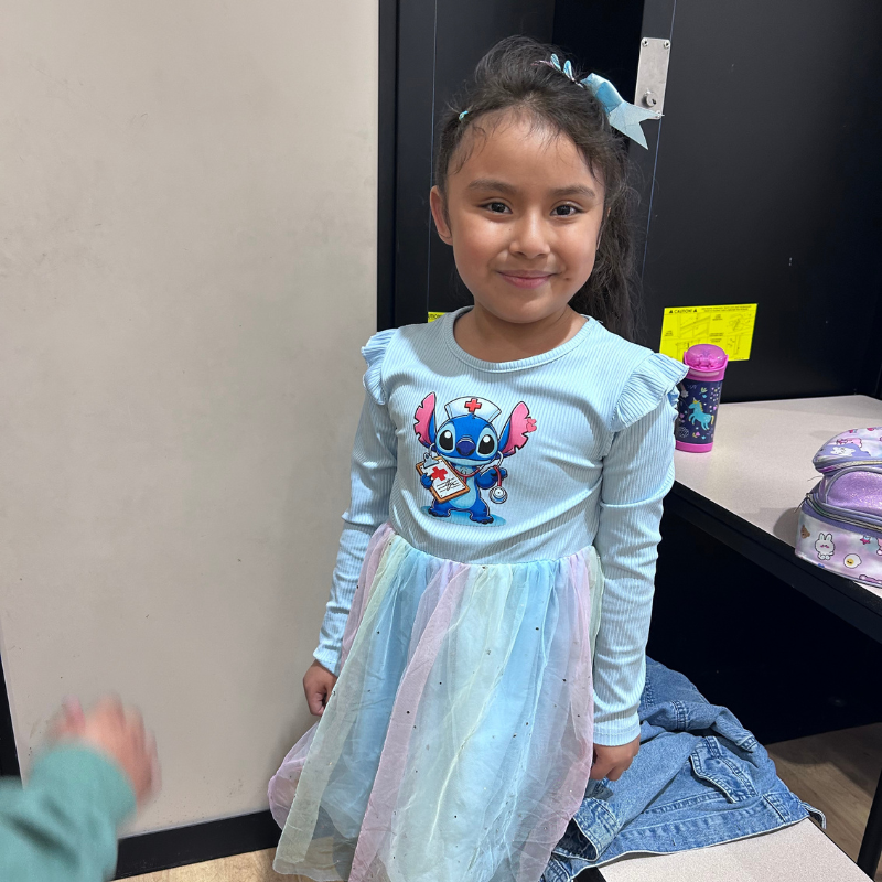 A young girl smiles, wearing a light blue long-sleeved shirt with a Stitch nurse graphic and a pastel rainbow tutu skirt.