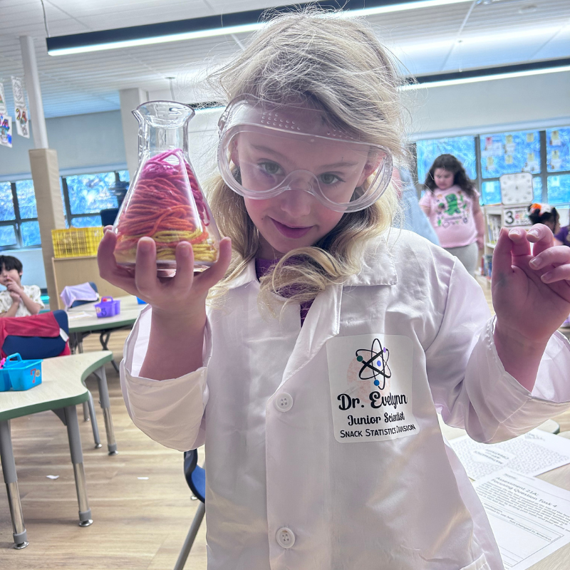 A young girl in a lab coat and goggles holds a flask filled with colorful yarn, engaged in a science experiment.