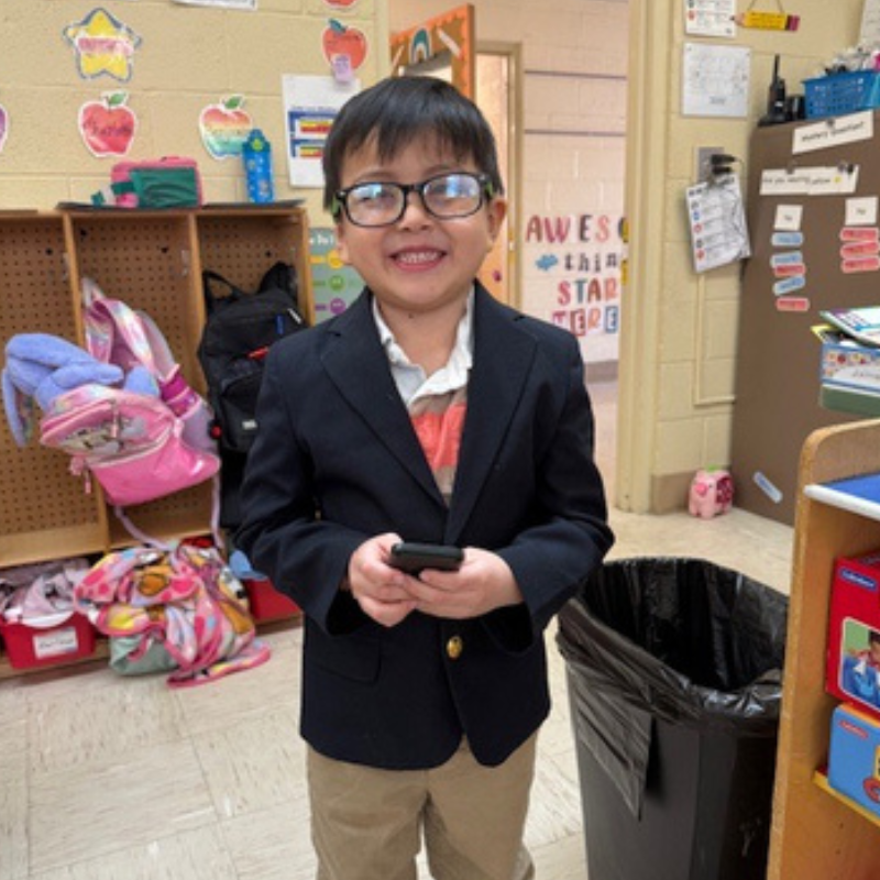 A young boy in a blazer and glasses smiles while holding a phone in a classroom.