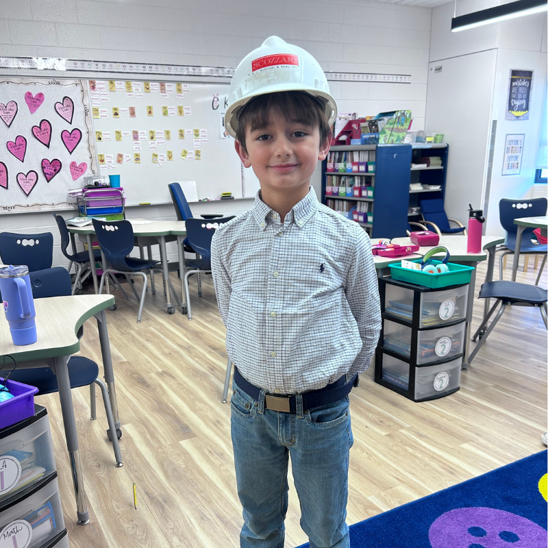 A young boy wearing a white hard hat and a plaid shirt stands in a classroom.