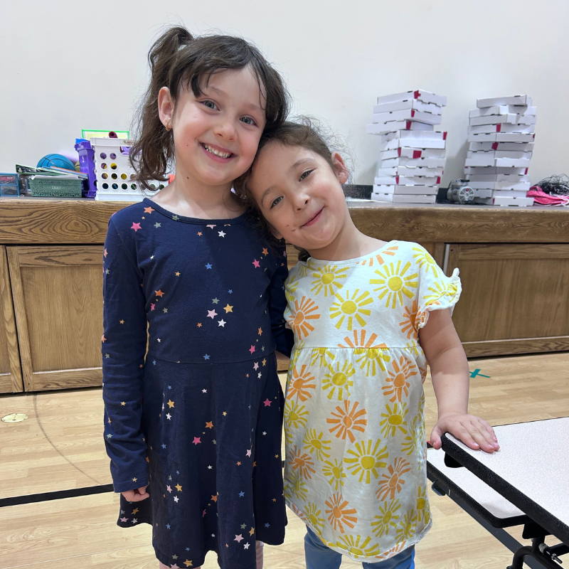 Two young girls smile at the camera, one with her head resting on the other's shoulder.